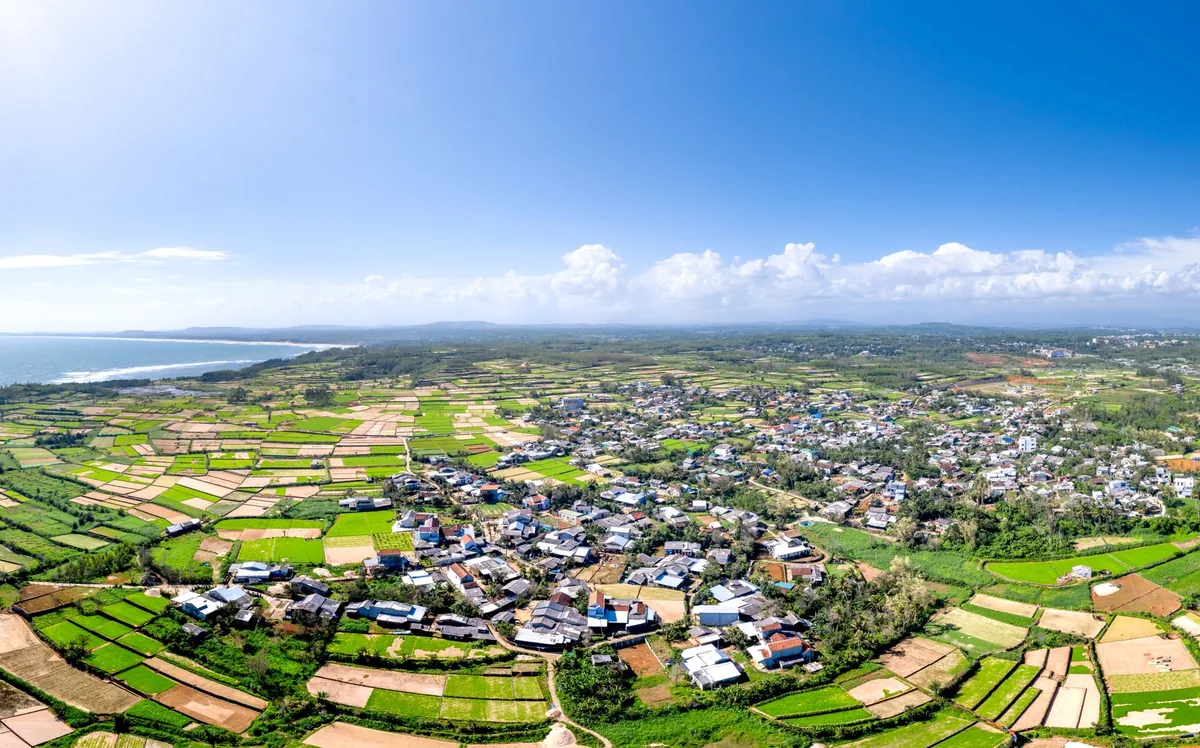 Vista aérea de um vilarejo rural cercado por plantações, sob céu azul.